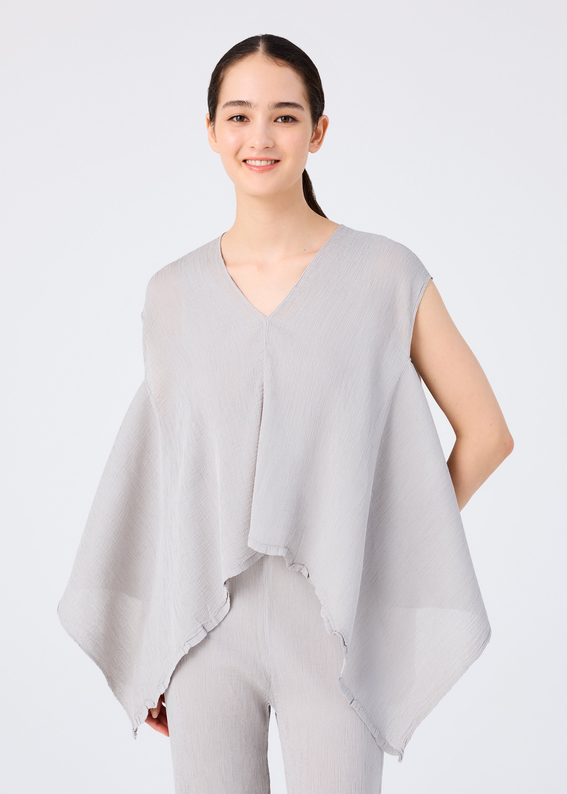 Zoomed view A young woman with long dark hair in a ponytail wears the me ISSEY MIYAKE FLAT SEE-THROUGH CREPE—a loose, sleeveless light gray top with wide sides over matching pants—standing against a plain white background and smiling gently.