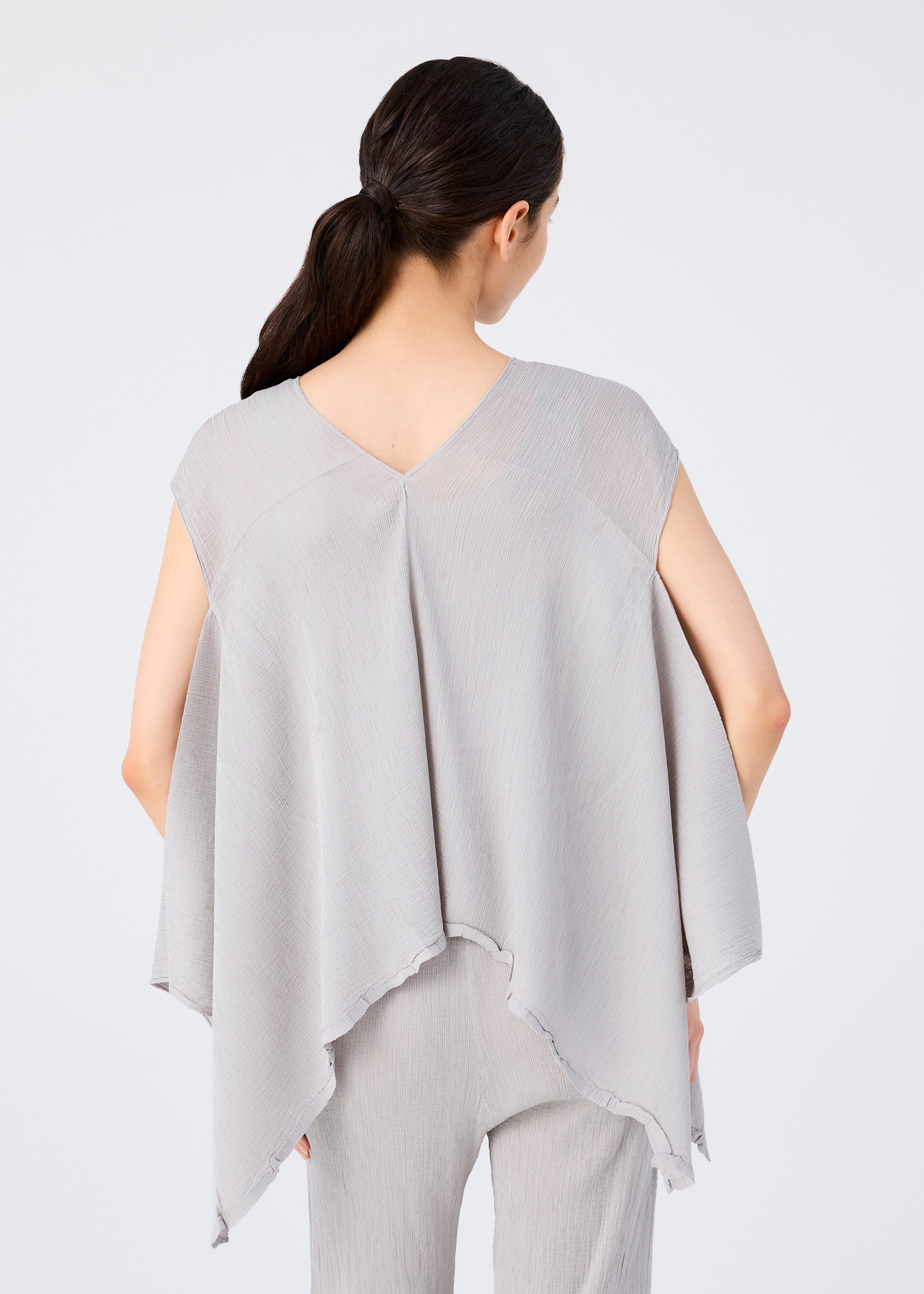 Zoomed view A woman with long dark hair in a low ponytail is seen from behind, wearing the me ISSEY MIYAKE FLAT SEE-THROUGH CREPE sleeveless top and matching pants, both featuring an uneven hem, against a plain light background.