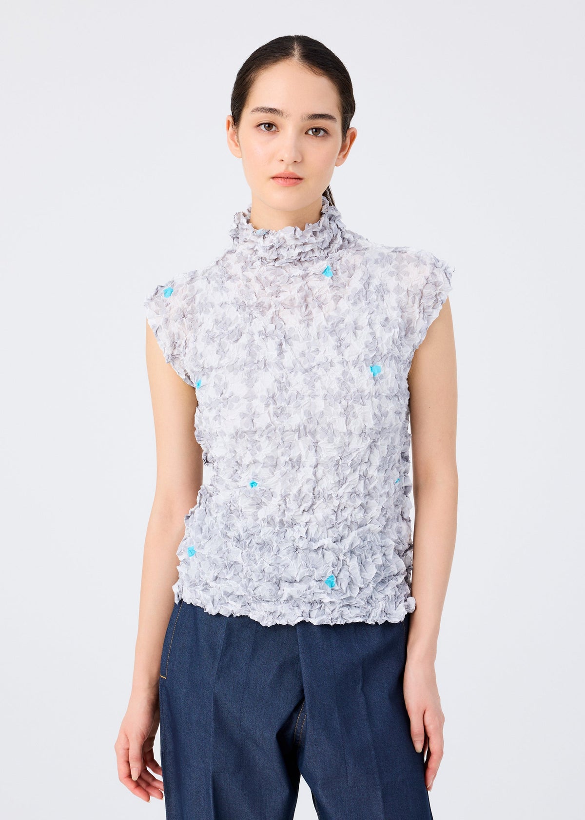 A woman with dark hair tied back models the SUIT MERINGUE TOP by me ISSEY MIYAKE—a high-neck sleeveless top with subtle playing card and blue details—paired with dark blue pants against a plain white background. view 6 of 8