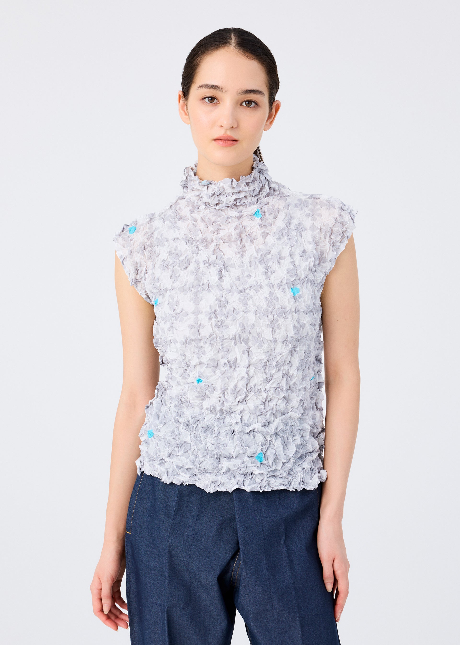 Zoomed view A woman with dark hair tied back models the SUIT MERINGUE TOP by me ISSEY MIYAKE—a high-neck sleeveless top with subtle playing card and blue details—paired with dark blue pants against a plain white background.