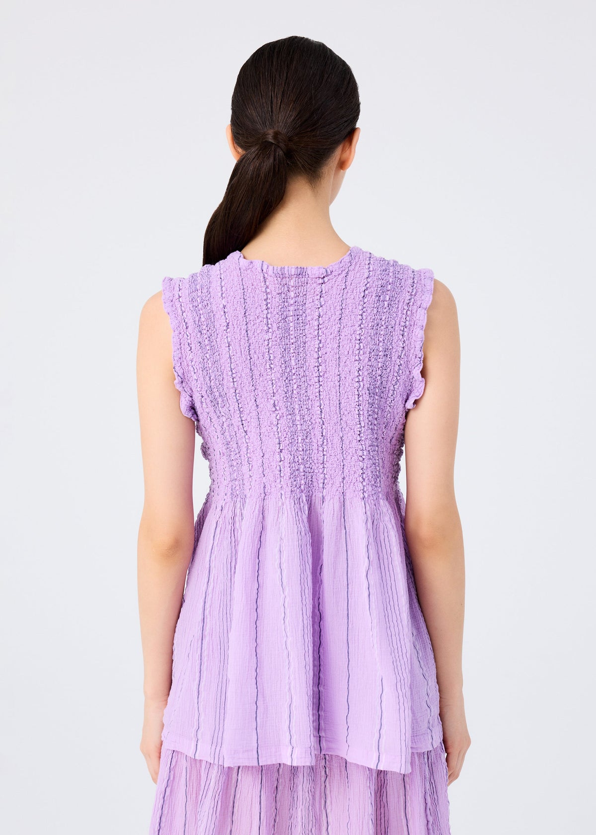 A woman with dark hair in a low ponytail is seen from the back, wearing the me ISSEY MIYAKE SHIRT STRIPE CAULIFLOWER, a sleeveless lavender top with textured cotton, flared hem, and ruffled armholes against a plain white background. view 6 of 7