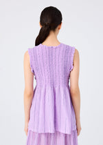 A woman with dark hair in a low ponytail is seen from the back, wearing the me ISSEY MIYAKE SHIRT STRIPE CAULIFLOWER, a sleeveless lavender top with textured cotton, flared hem, and ruffled armholes against a plain white background.