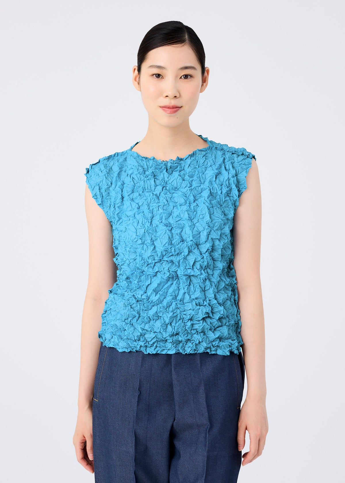 A woman with straight dark hair tied back wears the sleeveless MERINGUE TOP by me ISSEY MIYAKE in crushed pleat blue polyester and dark blue pants, standing facing forward against a plain white background. view 8 of 10