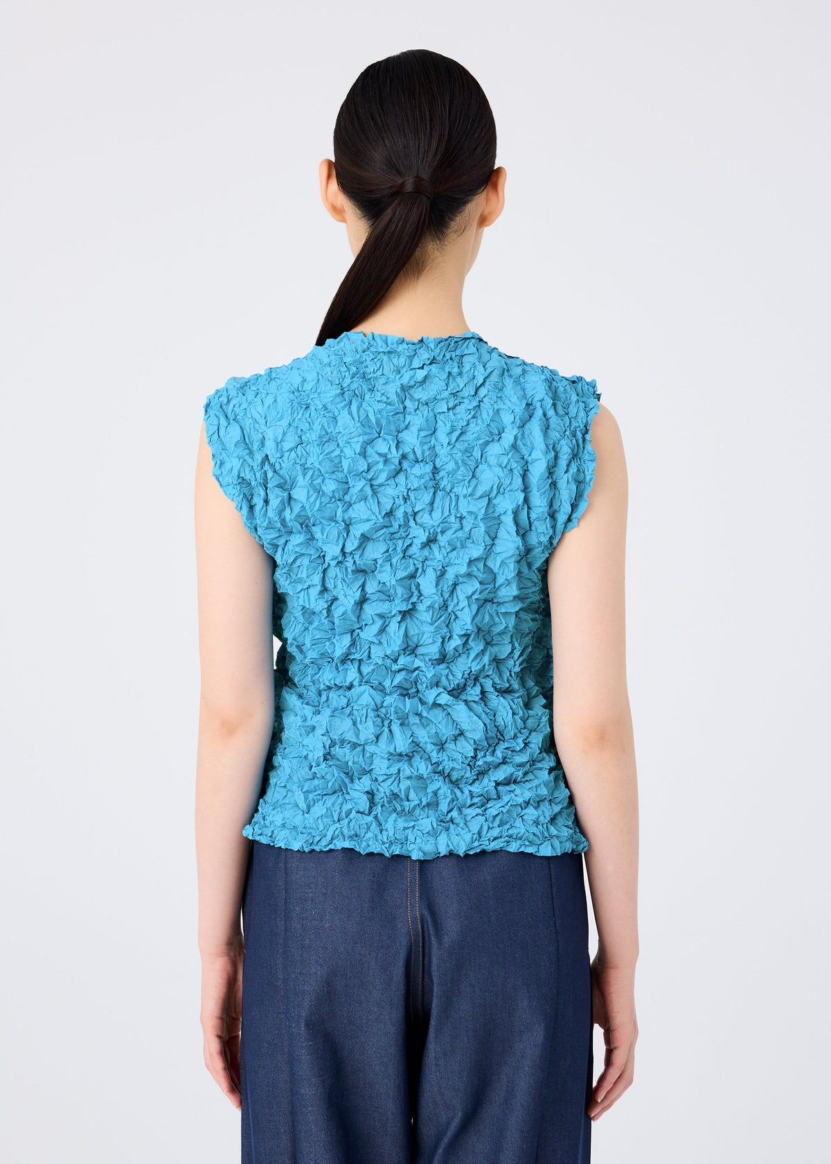 A person with long dark hair tied back is shown from behind wearing the MERINGUE TOP by me ISSEY MIYAKE—a blue, sleeveless, polyester crushed pleat top—paired with dark blue pants against a plain light background. view 9 of 10