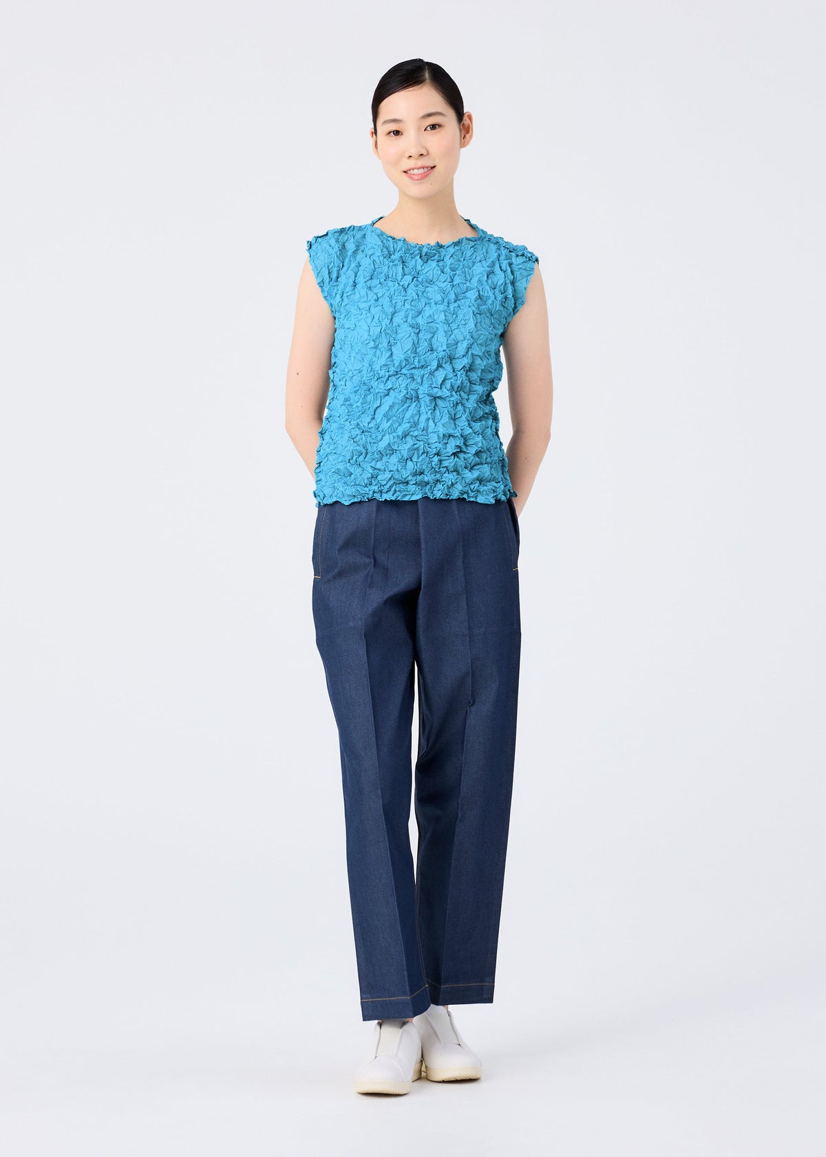 A person stands facing forward, smiling, wearing the MERINGUE TOP by me ISSEY MIYAKE—a textured blue sleeveless polyester top—paired with dark blue pants and white shoes, hands behind their back, against a plain white background. view 10 of 10