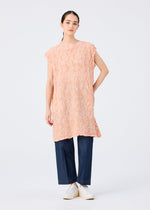 A woman with straight dark hair pulled back and a neutral expression wears the me ISSEY MIYAKE SURPRISE BOX PRINT CAULIFLOWER, a loose peach cotton tunic with subtle pigment printing, styled over dark wide-leg jeans and white slip-on shoes against a plain white background.