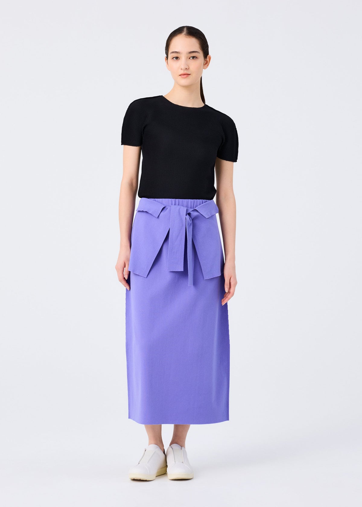A woman stands against a plain background wearing the FLAT JERSEY top by me ISSEY MIYAKE, paired with a high-waisted lavender cotton-blend skirt featuring a large waist bow, and white slip-on shoes. view 6 of 8