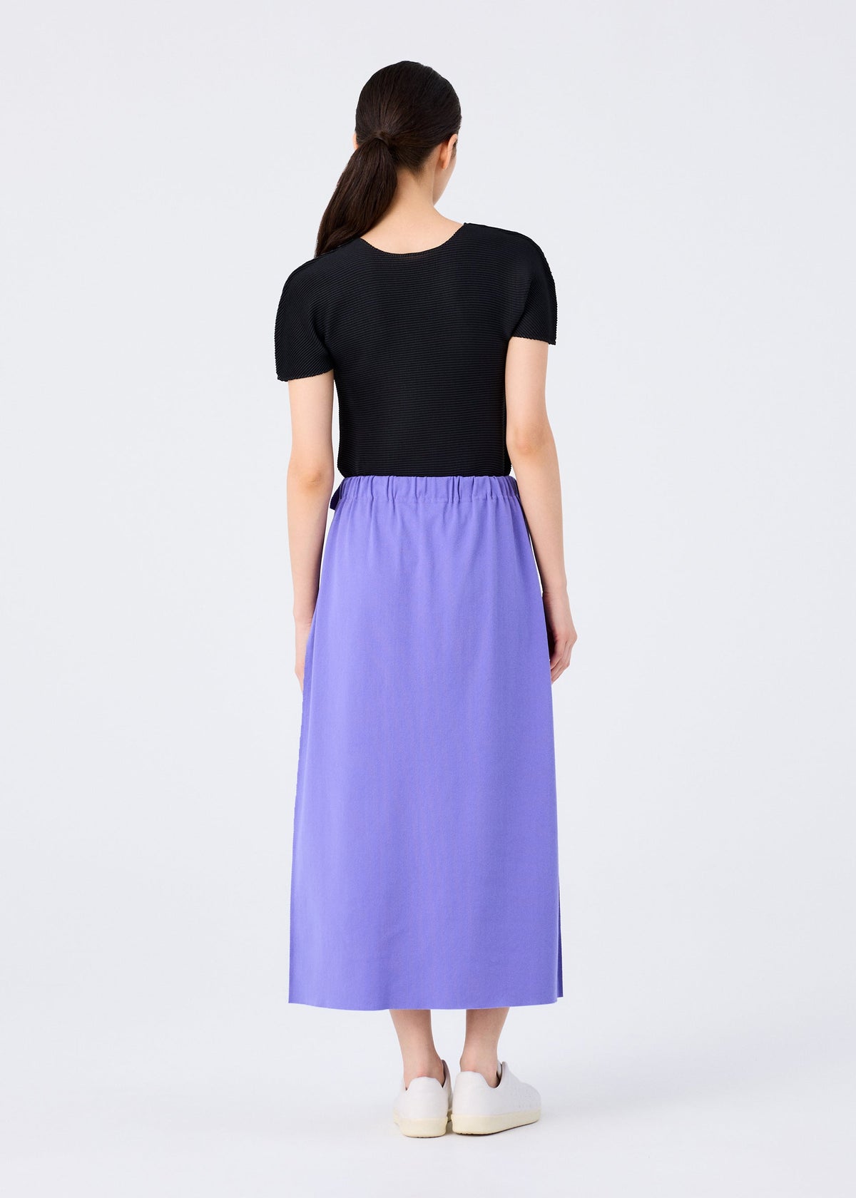 A woman with long dark hair pulled back models the FLAT JERSEY by me ISSEY MIYAKE—a short-sleeved black top and high-waisted, ankle-length versatile skirt—paired with white slip-on shoes against a plain light background. view 7 of 8