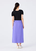 A woman with long dark hair pulled back models the FLAT JERSEY by me ISSEY MIYAKE—a short-sleeved black top and high-waisted, ankle-length versatile skirt—paired with white slip-on shoes against a plain light background.