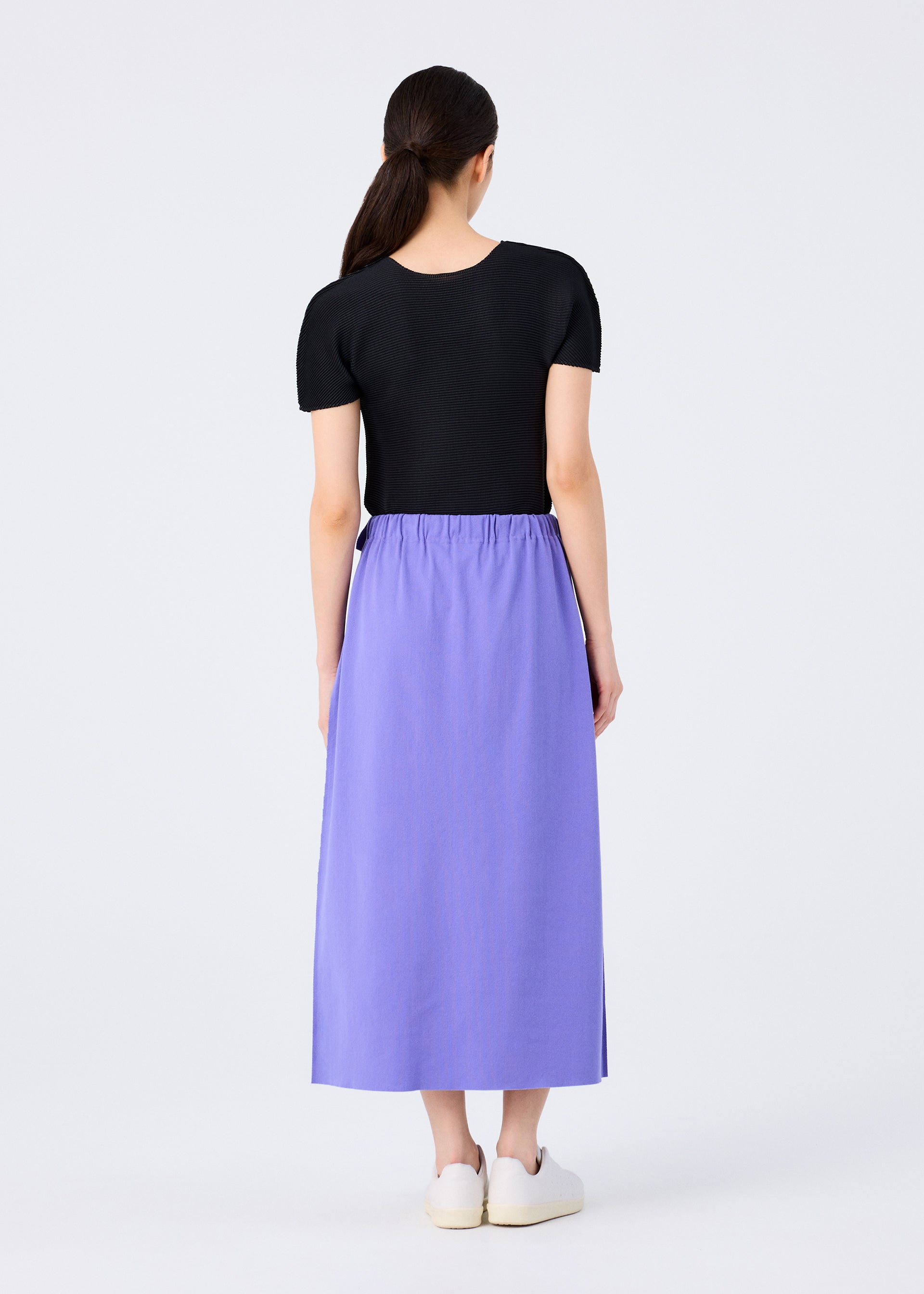 Zoomed view A woman with long dark hair pulled back models the FLAT JERSEY by me ISSEY MIYAKE—a short-sleeved black top and high-waisted, ankle-length versatile skirt—paired with white slip-on shoes against a plain light background.