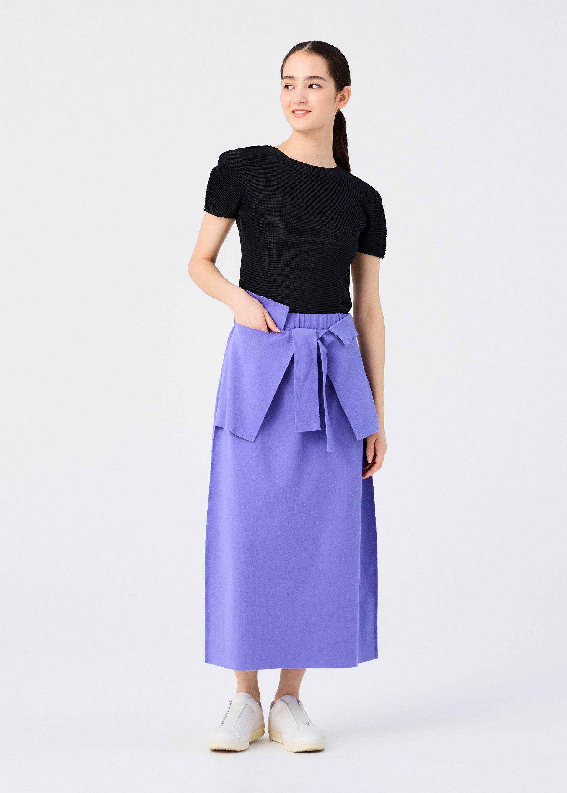 Zoomed view A woman smiles slightly and looks to the side while wearing the FLAT JERSEY short-sleeved black top from me ISSEY MIYAKE, paired with a long cotton-blend skirt, wide tied belt, and white slip-on shoes against a white background.