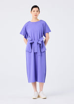 A woman with short dark hair stands facing forward in a loose-fitting, short-sleeved FLAT JERSEY dress by me ISSEY MIYAKE. The lavender cotton-blend dress features a wide matching belt and is paired with white slip-on shoes against a plain background.