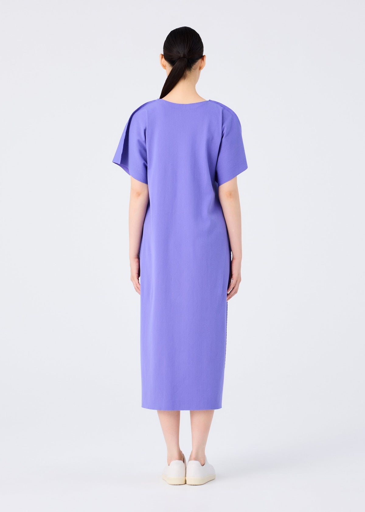 A person with long dark hair in a low ponytail faces away, wearing the me ISSEY MIYAKE FLAT JERSEY loose-fitting, short-sleeved, knee-length cotton-blend dress in purple, paired with white shoes against a plain white background. view 7 of 8