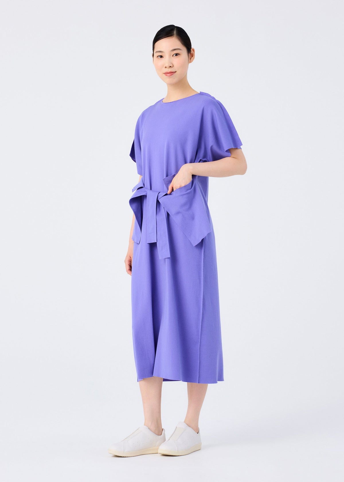 A person stands against a plain white background, wearing the FLAT JERSEY dress by me ISSEY MIYAKE—a loose-fitting, knee-length purple cotton-blend dress with short sleeves and a tied waist—paired with white slip-on shoes. view 8 of 8