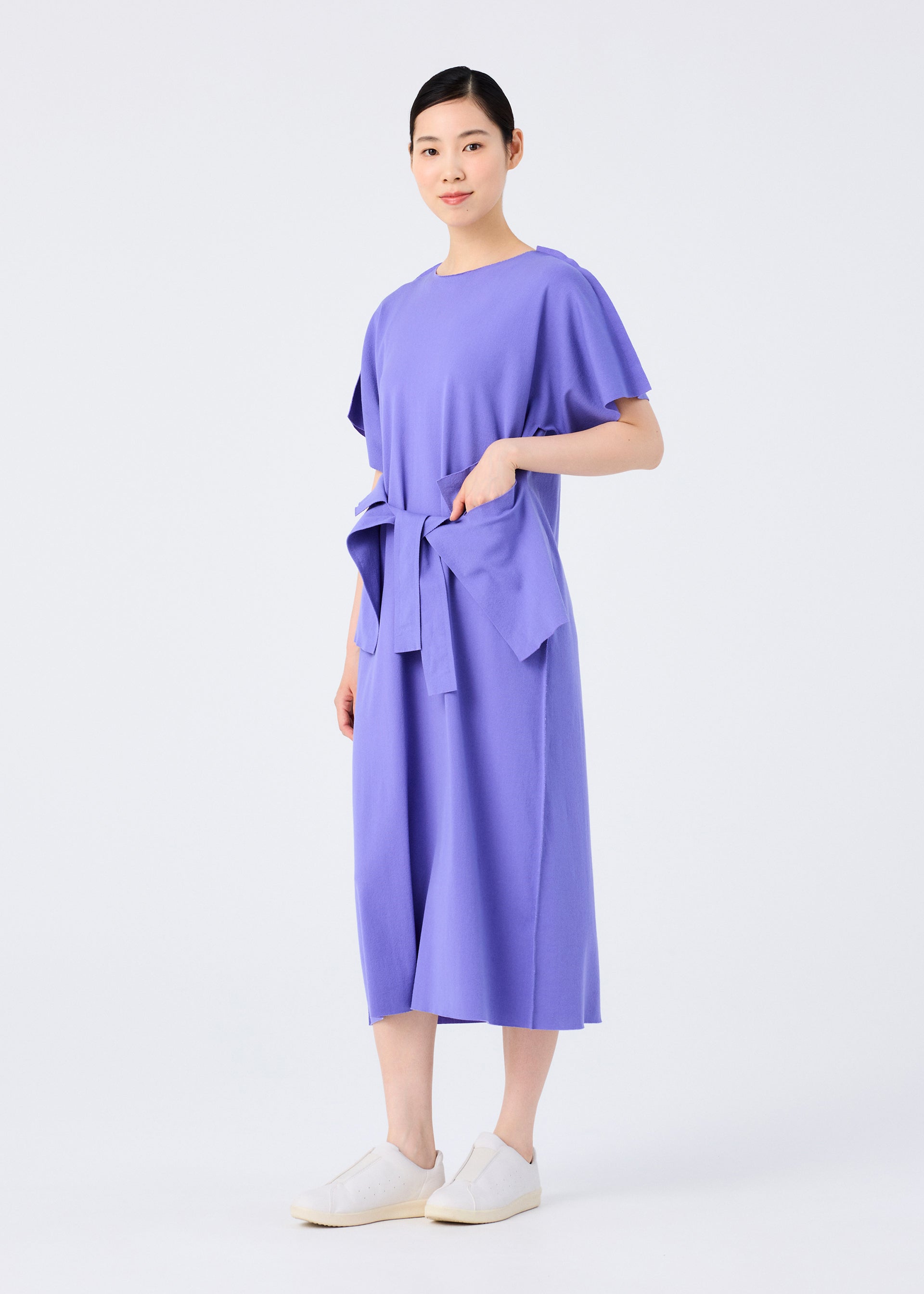 Zoomed view A person stands against a plain white background, wearing the FLAT JERSEY dress by me ISSEY MIYAKE—a loose-fitting, knee-length purple cotton-blend dress with short sleeves and a tied waist—paired with white slip-on shoes.