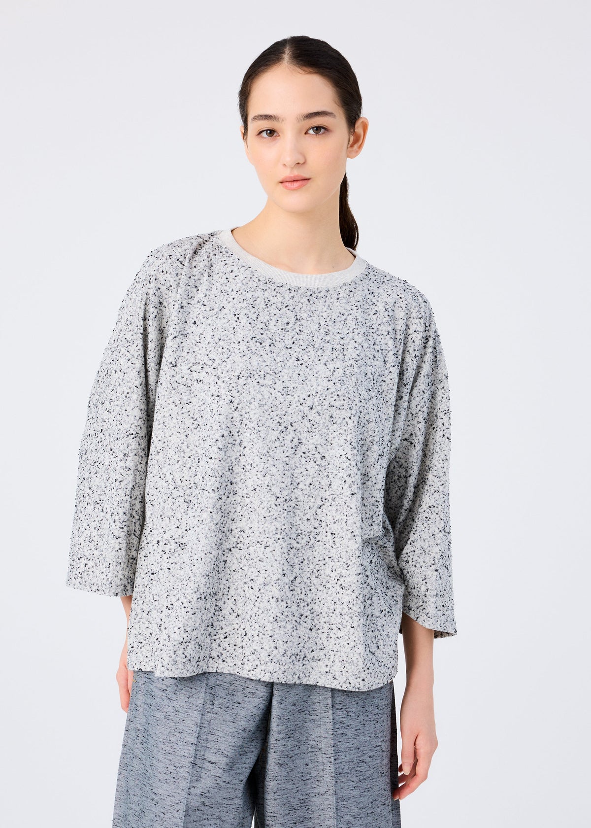 A woman with long dark hair tied back wears the me ISSEY MIYAKE REmeTEX NEP JERSEY—a loose-fitting, light gray speckled top with three-quarter sleeves and matching gray pants—made from recycled polyester, standing against a plain white background. view 3 of 5