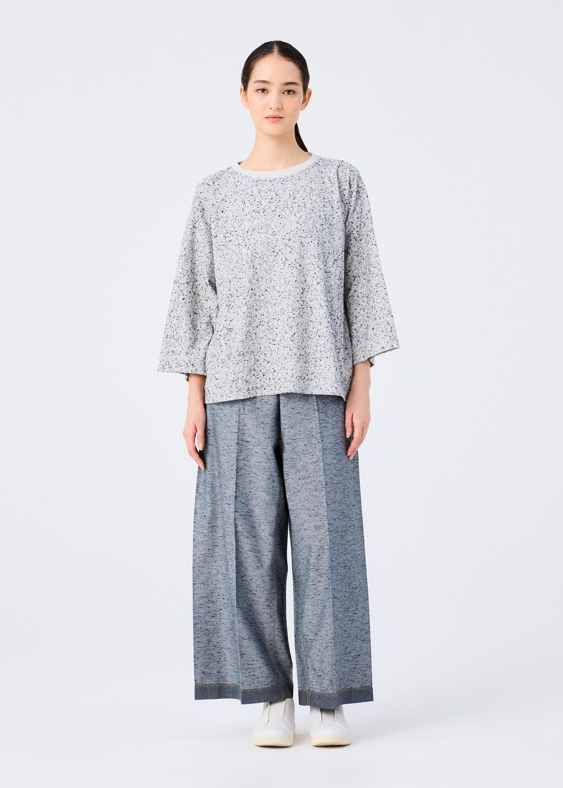 Zoomed view A woman with straight dark hair pulled back wears a me ISSEY MIYAKE REmeTEX NEP JERSEY light gray patterned top with wide sleeves and light blue-gray wide-leg pants, paired with white shoes, all set against a plain white background.