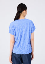 A person with dark hair tied back faces away, wearing the light blue MERINGUE T-SHIRT by me ISSEY MIYAKE paired with dark pants, set against a plain white background.