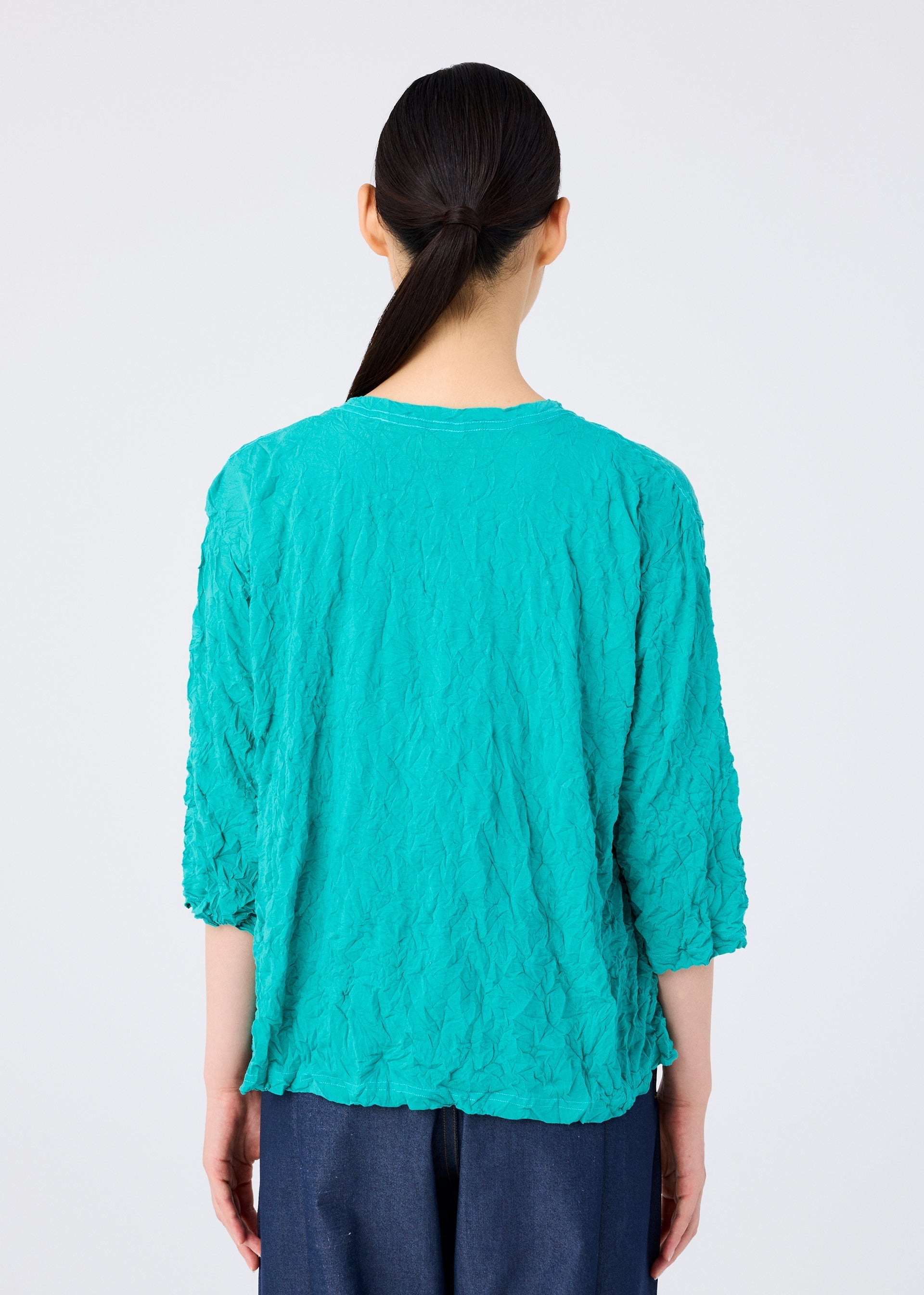 Zoomed view A person with long dark hair in a low ponytail is seen from behind, wearing the MERINGUE T-SHIRT by me ISSEY MIYAKE in textured turquoise with three-quarter sleeves and dark pants, against a plain light background.