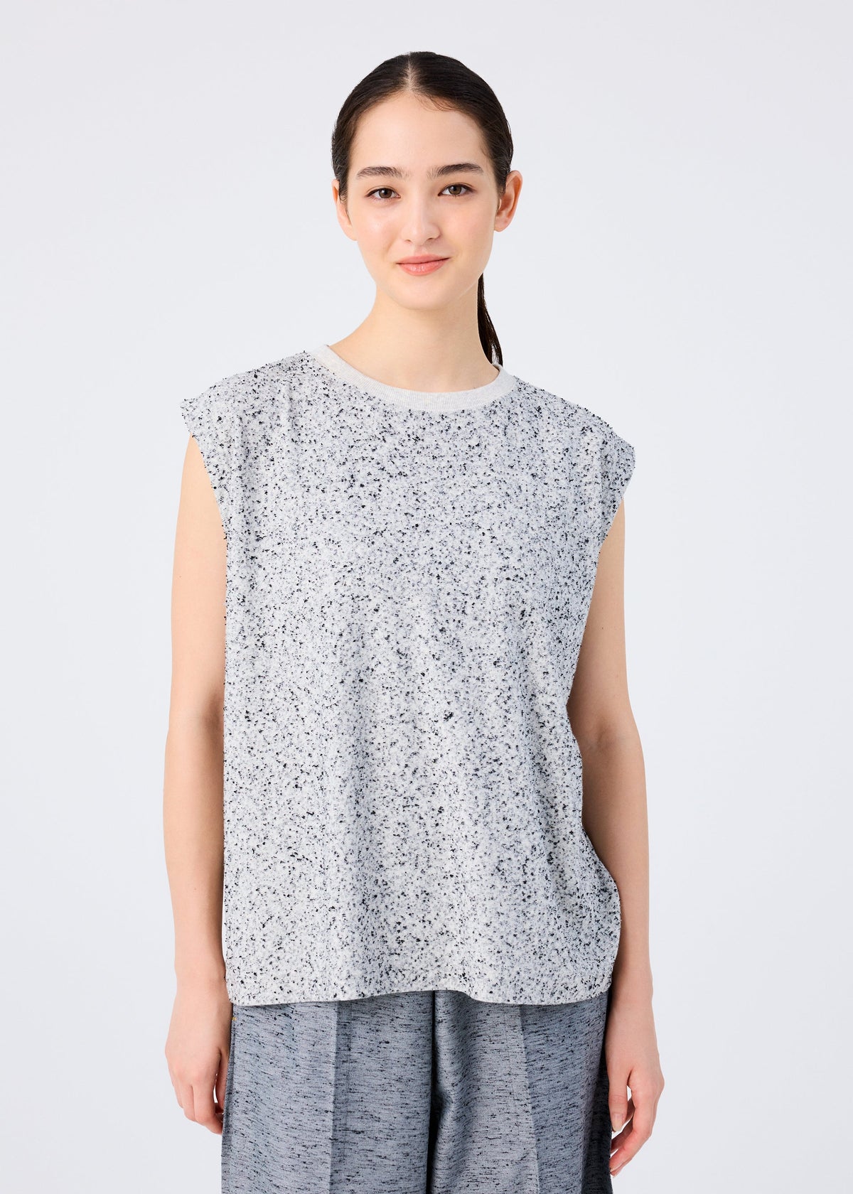 A young woman with long dark hair pulled back smiles gently at the camera, wearing the me ISSEY MIYAKE REmeTEX NEP JERSEY sleeveless top and loose gray pants, standing against a plain white background. view 3 of 5