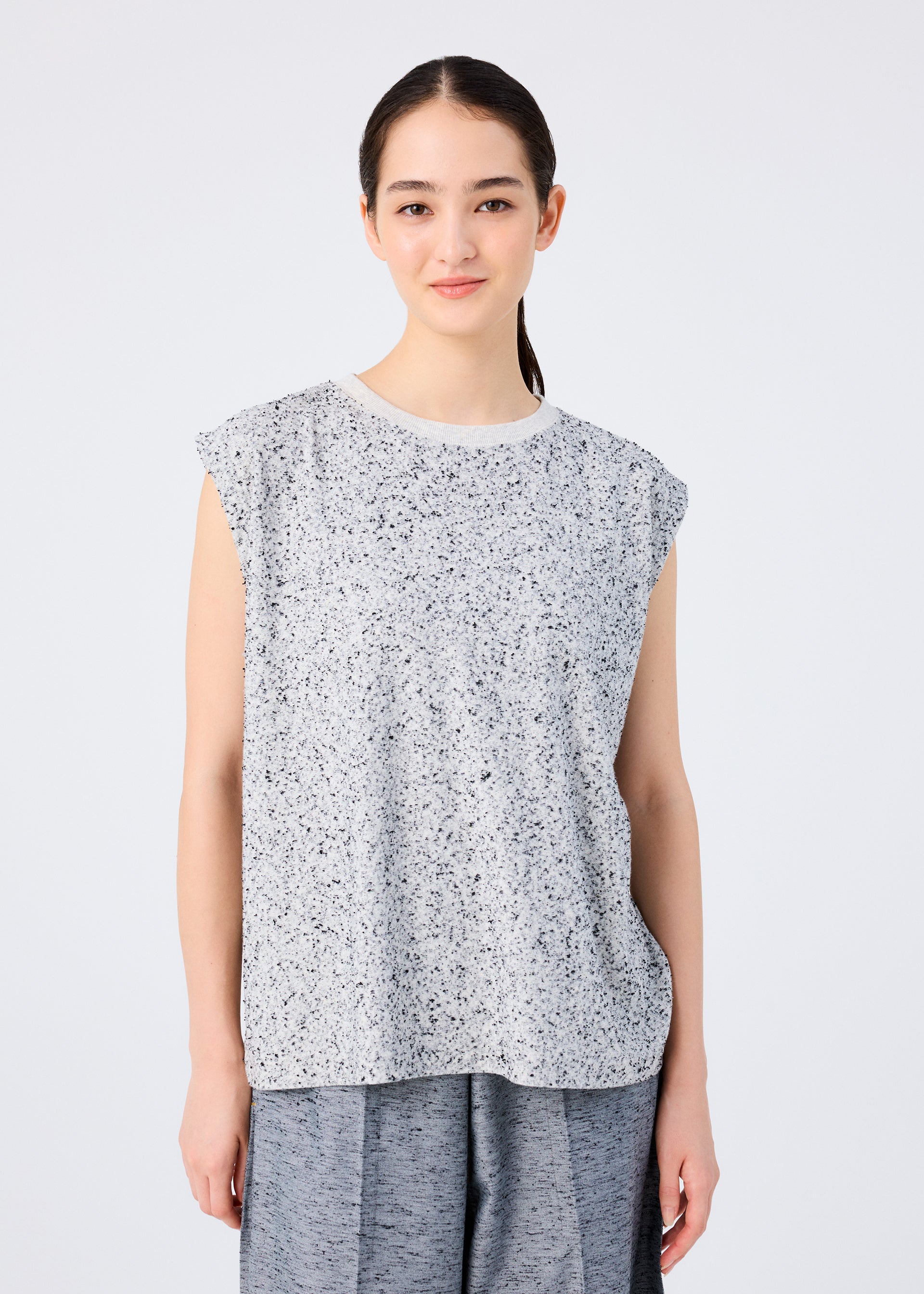 Zoomed view A young woman with long dark hair pulled back smiles gently at the camera, wearing the me ISSEY MIYAKE REmeTEX NEP JERSEY sleeveless top and loose gray pants, standing against a plain white background.