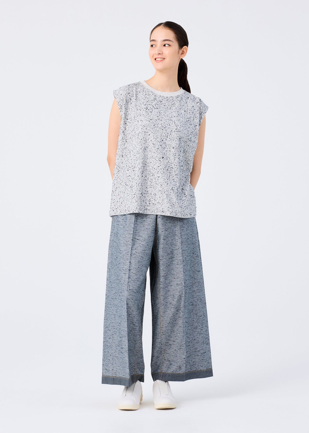 A woman stands against a plain background, smiling. She wears the me ISSEY MIYAKE REmeTEX NEP JERSEY sleeveless top with wide-leg striped gray pants and white shoes. Her hair is tied back in a ponytail. view 5 of 5