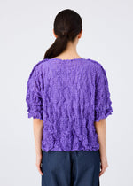 A woman with long dark hair in a low ponytail is shown from the back, wearing the me ISSEY MIYAKE KALEIDOSCOPE LACE MERINGUE top in purple with short sleeves and dark blue pants against a plain light background.