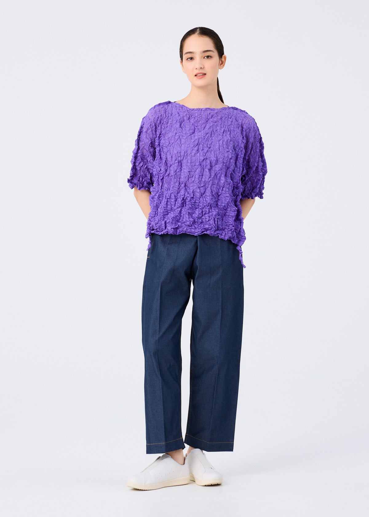 A person stands against a plain white background, wearing the KALEIDOSCOPE LACE MERINGUE top by me ISSEY MIYAKE in purple, paired with loose dark blue pants and white shoes, with their hands behind their back. view 10 of 10