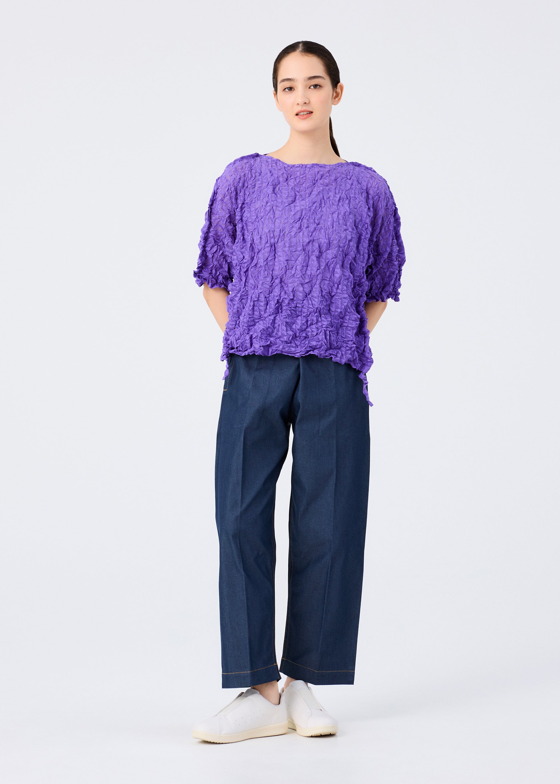 Zoomed view A person stands against a plain white background, wearing the KALEIDOSCOPE LACE MERINGUE top by me ISSEY MIYAKE in purple, paired with loose dark blue pants and white shoes, with their hands behind their back.