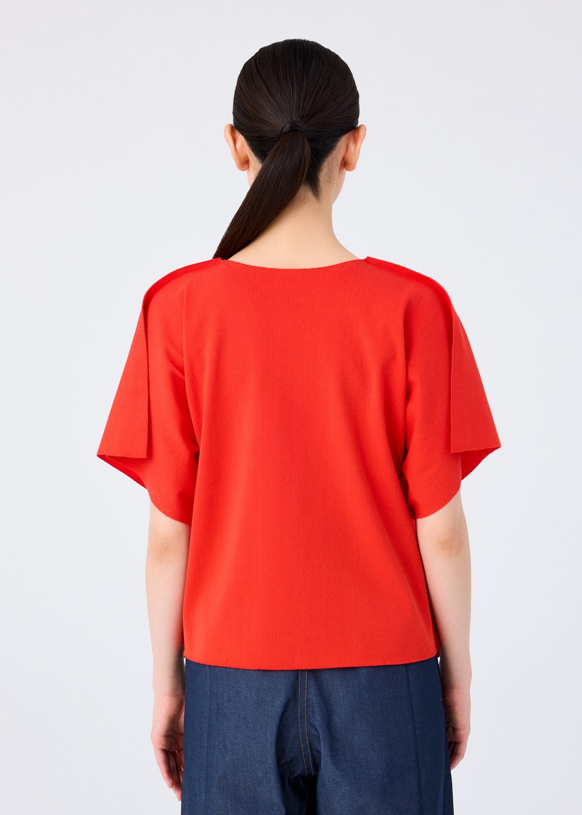 A person with long dark hair in a low ponytail is seen from the back, wearing the bright red FLAT JERSEY short-sleeved top by me ISSEY MIYAKE and dark blue pants, standing against a plain light gray background. view 7 of 8