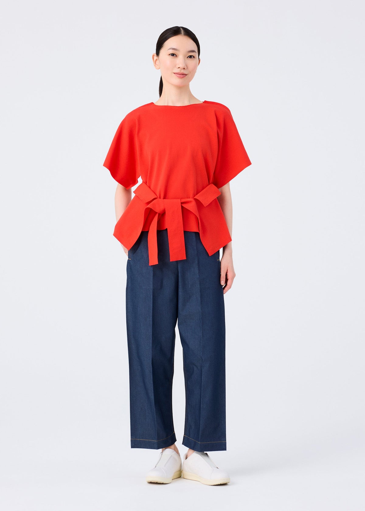 A woman with straight, pulled-back dark hair and a neutral expression wears the me ISSEY MIYAKE FLAT JERSEY—a bright red, cotton-blend short-sleeved top with a wide waist tie—paired with loose dark blue pants and white slip-on shoes. view 8 of 8