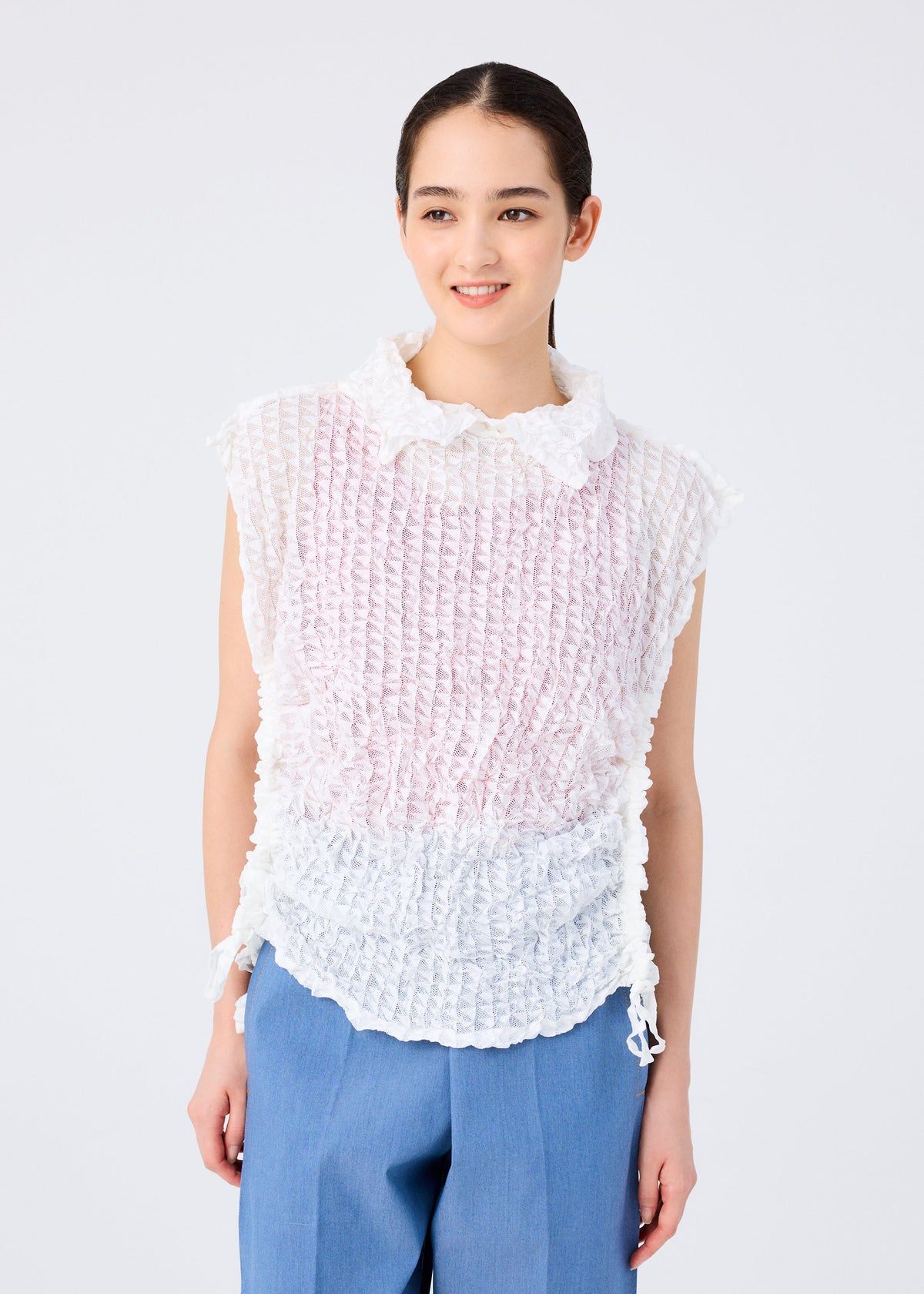 A woman with dark hair pulled back smiles, wearing the sleeveless KALEIDOSCOPE LACE MERINGUE top in white and pale pink crushed pleat by me ISSEY MIYAKE, paired with light blue trousers against a plain white background. view 8 of 10