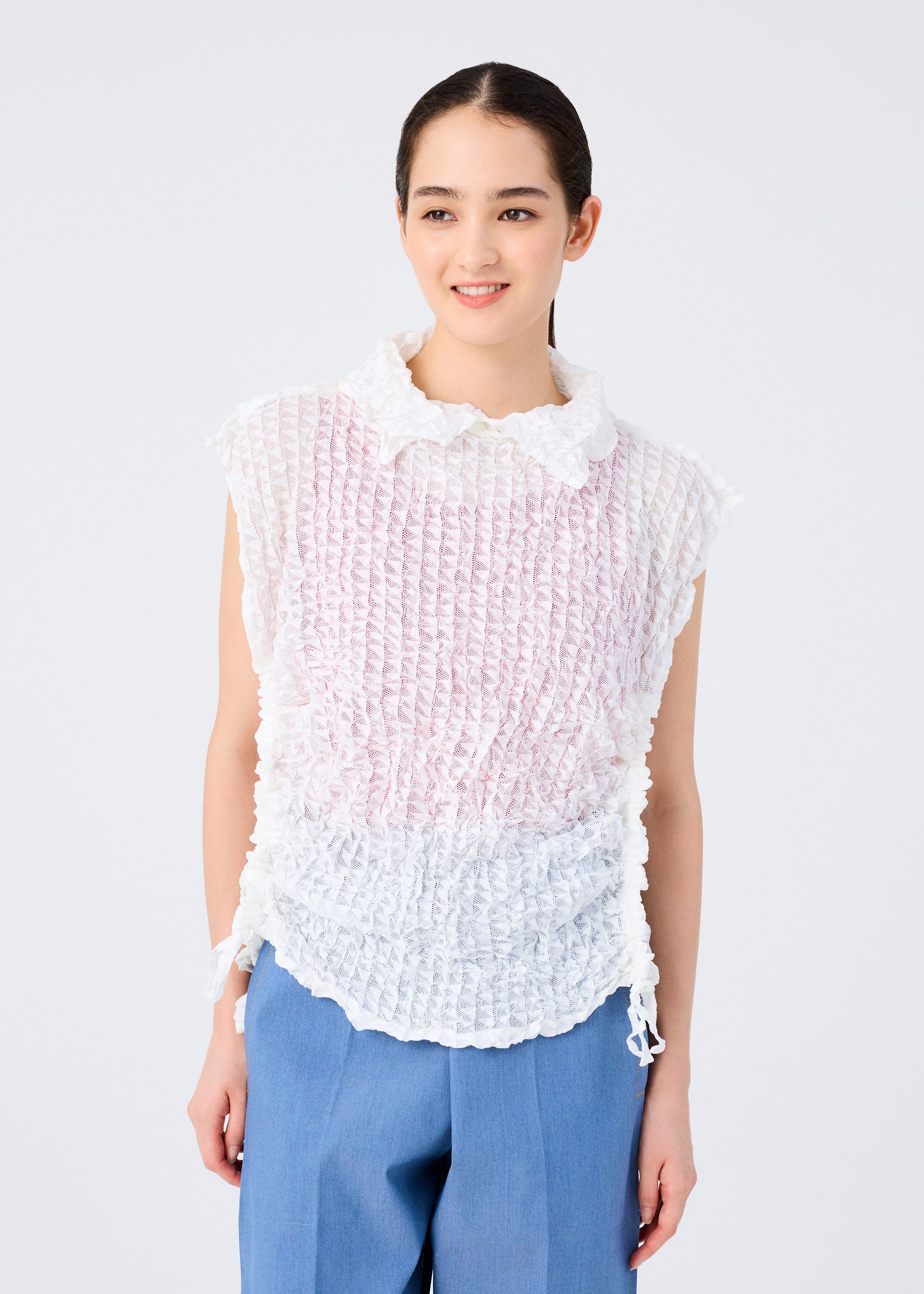 Zoomed view A woman with dark hair pulled back smiles, wearing the sleeveless KALEIDOSCOPE LACE MERINGUE top in white and pale pink crushed pleat by me ISSEY MIYAKE, paired with light blue trousers against a plain white background.