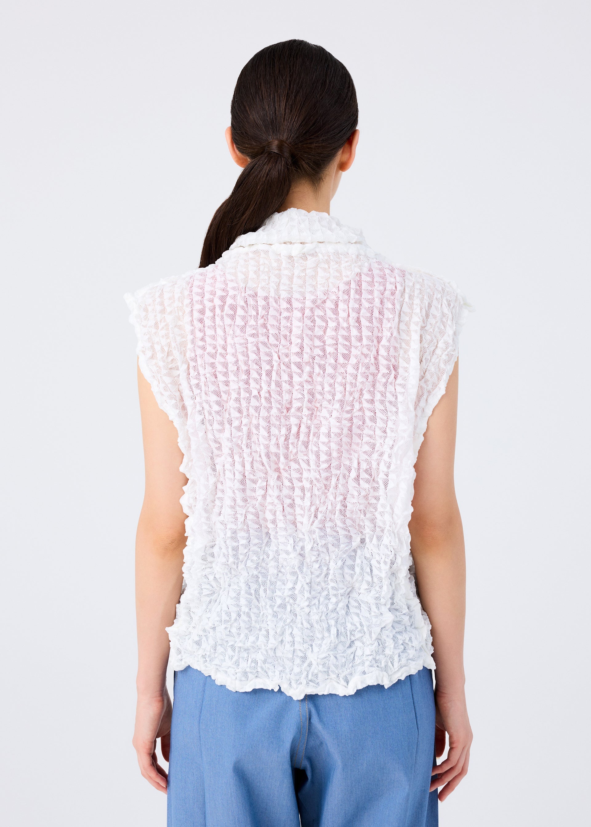 Zoomed view A person with long dark hair in a low ponytail is shown from the back, wearing the KALEIDOSCOPE LACE MERINGUE top by me ISSEY MIYAKE and blue pants against a plain white background.