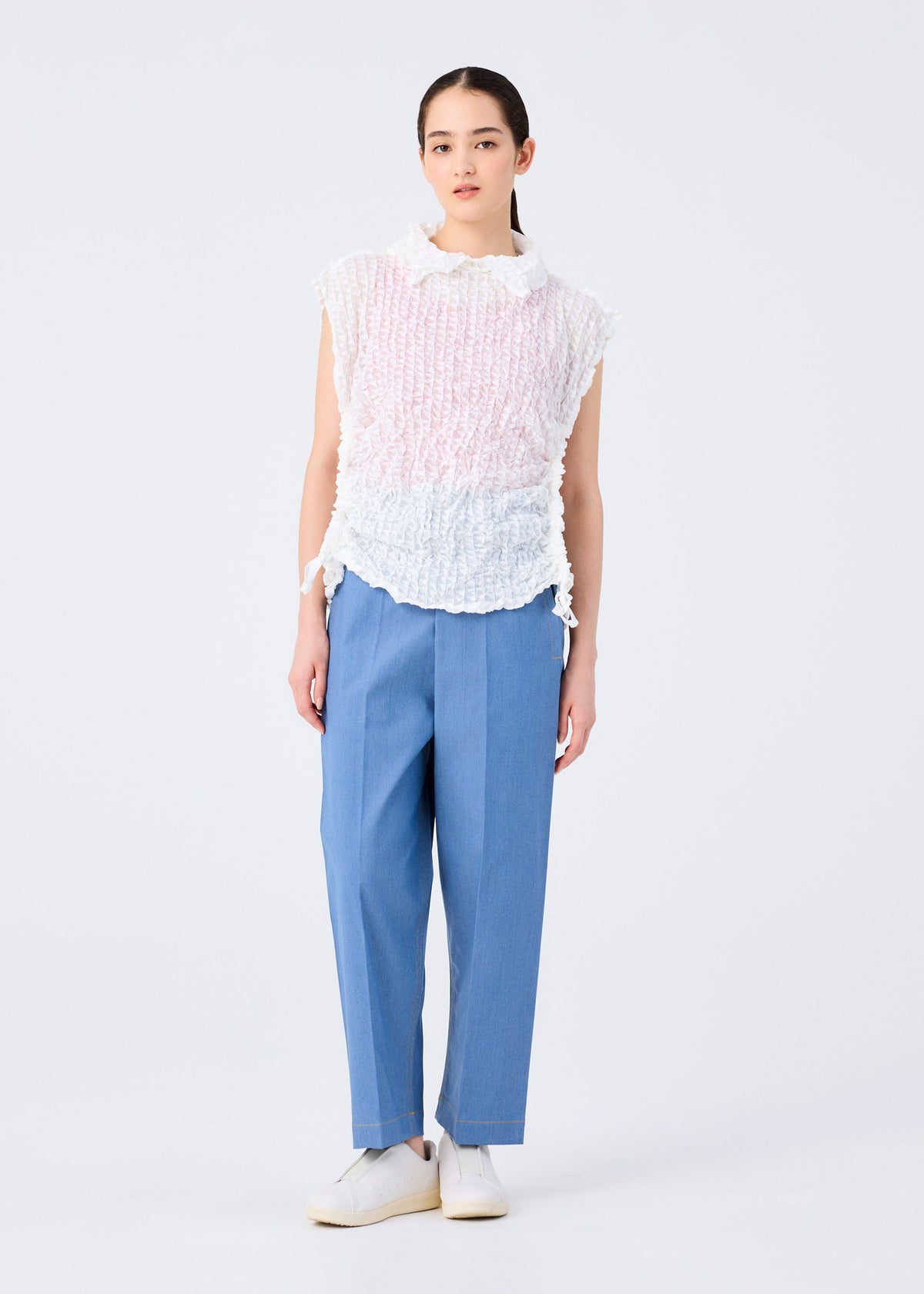 A woman with straight dark hair pulled back wears the KALEIDOSCOPE LACE MERINGUE sleeveless top by me ISSEY MIYAKE in white and pale pink, paired with blue high-waisted trousers and white shoes against a plain white background. view 10 of 10