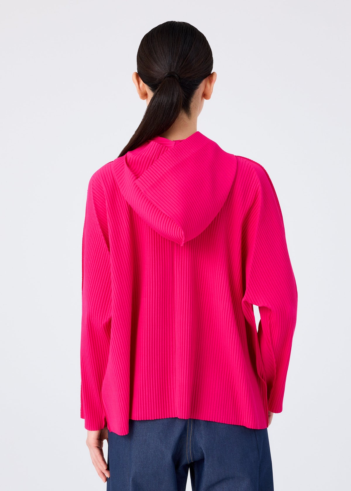 A person with long dark hair in a low ponytail is shown from the back, wearing the FINE KNIT PLEATS COLOR 1 hoodie by me ISSEY MIYAKE and dark blue pants against a plain light background. view 8 of 9