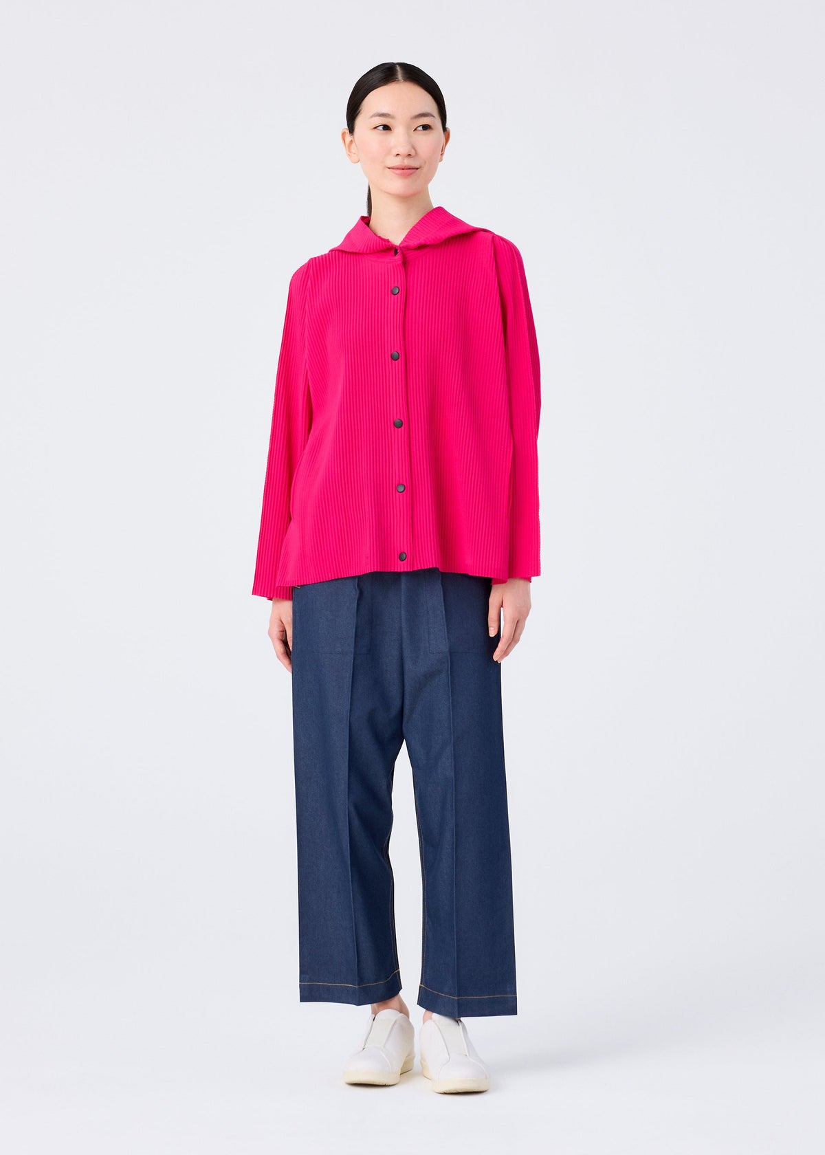 A woman wearing the me ISSEY MIYAKE FINE KNIT PLEATS COLOR 1 top stands against a white background. The bright pink, pleated, collared, long-sleeve button-up is paired with wide-leg dark blue pants and white shoes as she smiles gently. view 9 of 9