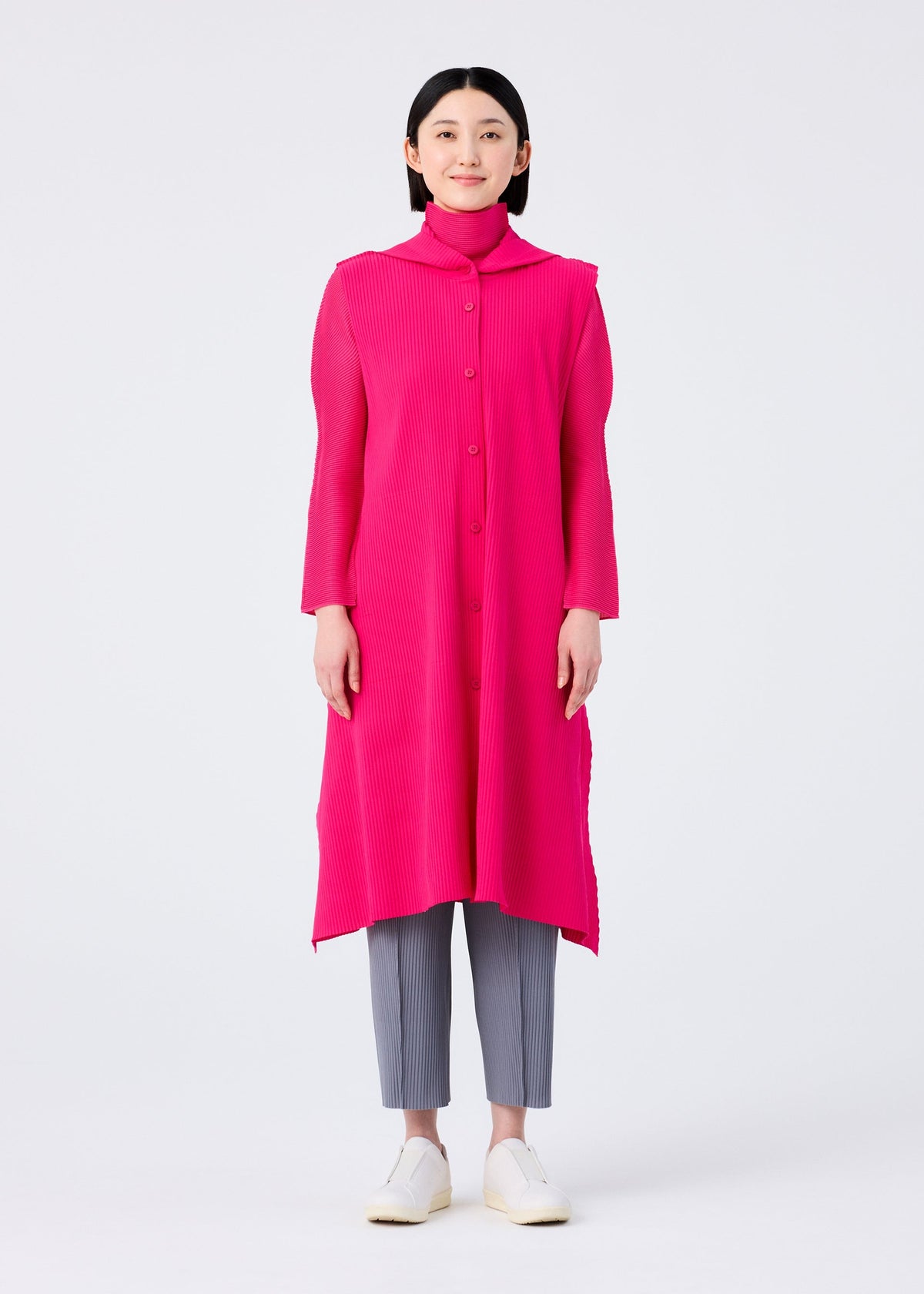 A woman faces forward against a white background, wearing the FLARE PLEATS long bright pink vest by me ISSEY MIYAKE over a matching turtleneck, paired with gray cropped pants and white slip-on shoes. view 6 of 8