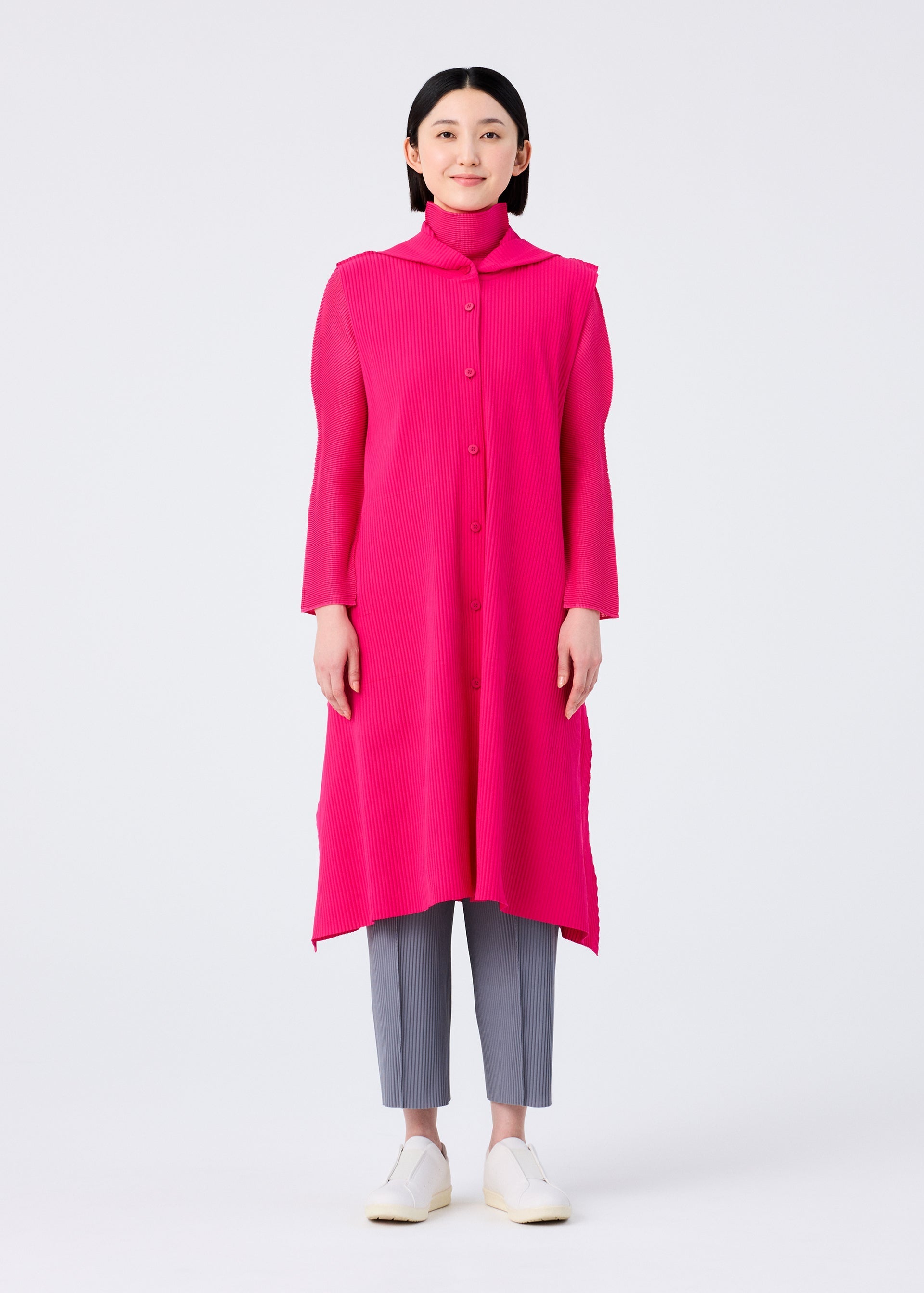 Zoomed view A woman faces forward against a white background, wearing the FLARE PLEATS long bright pink vest by me ISSEY MIYAKE over a matching turtleneck, paired with gray cropped pants and white slip-on shoes.