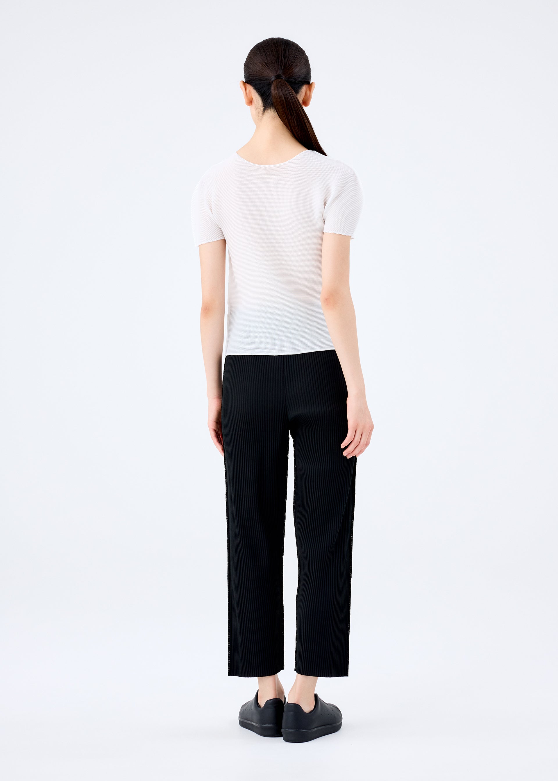 Zoomed view A woman with long dark hair in a low ponytail stands facing away, wearing me ISSEY MIYAKE FINE KNIT PLEATS BLACK pants, a short-sleeved white top, and black shoes against a plain white background.