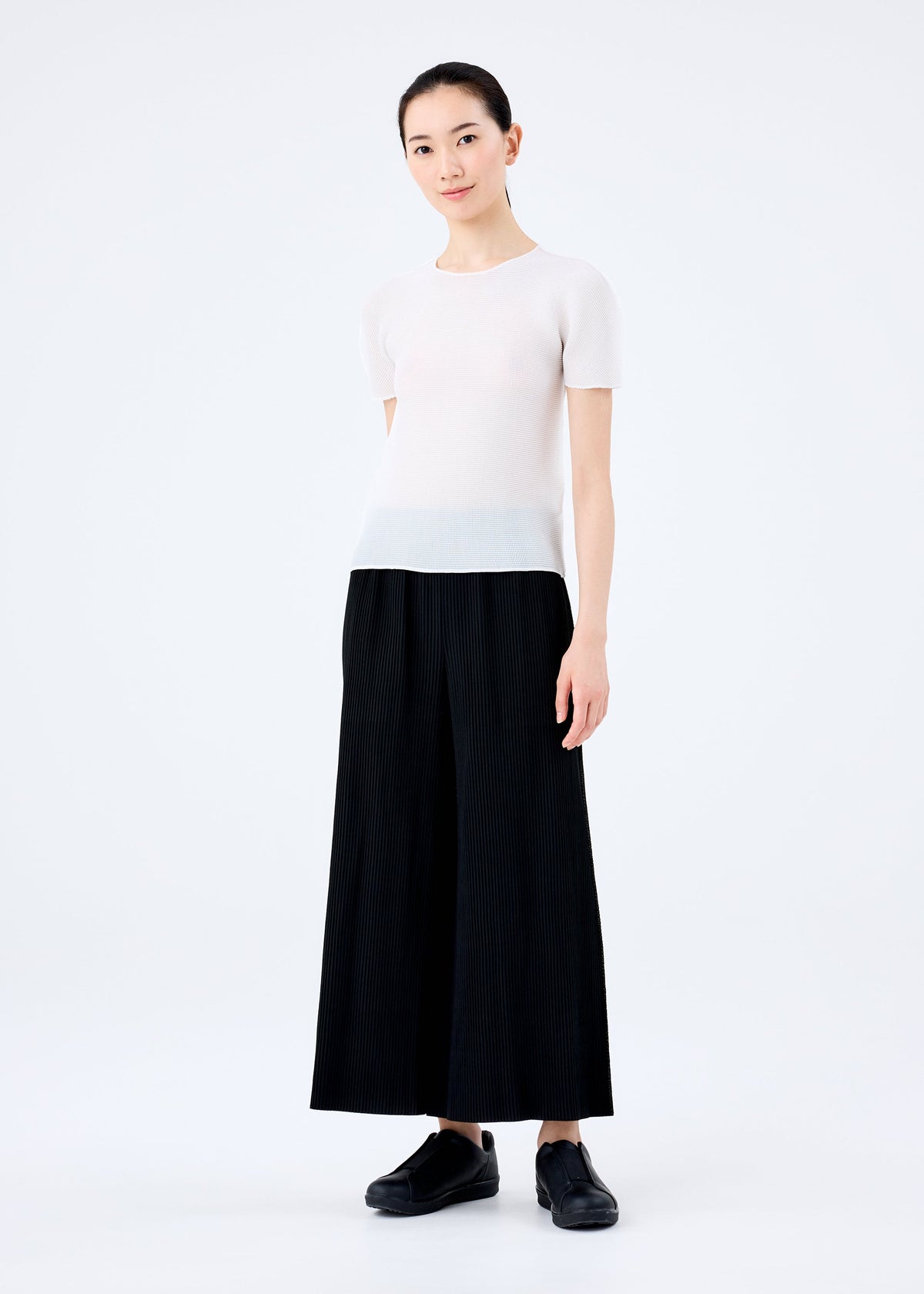 A woman with her hair tied back smiles gently at the camera, wearing the me ISSEY MIYAKE FINE KNIT PLEATS BLACK pants, a short-sleeved white top, and black shoes against a plain white background. view 3 of 5