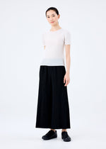 A woman with her hair tied back smiles gently at the camera, wearing the me ISSEY MIYAKE FINE KNIT PLEATS BLACK pants, a short-sleeved white top, and black shoes against a plain white background.