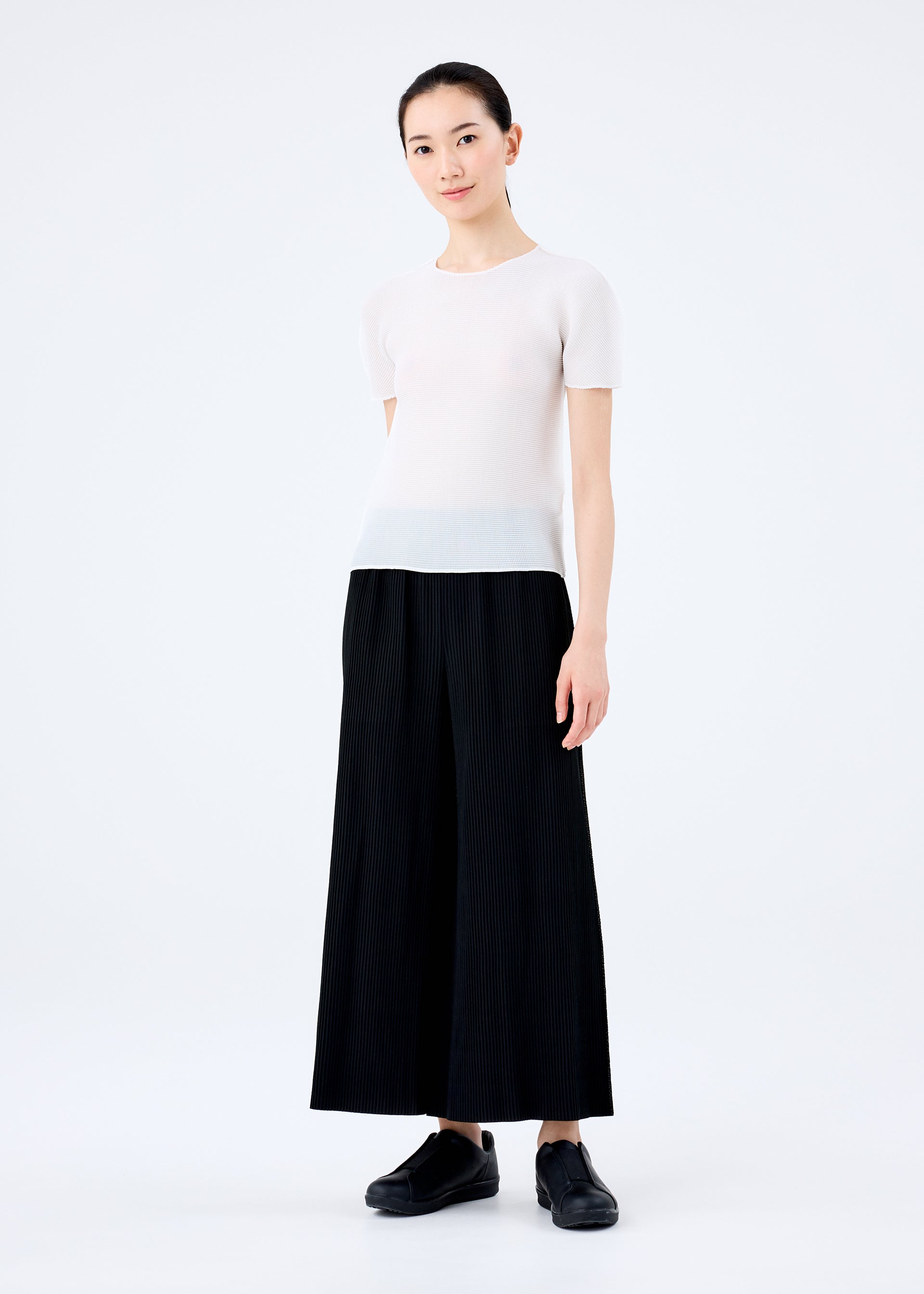 Zoomed view A woman with her hair tied back smiles gently at the camera, wearing the me ISSEY MIYAKE FINE KNIT PLEATS BLACK pants, a short-sleeved white top, and black shoes against a plain white background.