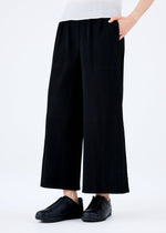 A person wears the FINE KNIT PLEATS BLACK pants by me ISSEY MIYAKE with their right hand in a pocket, paired with black slip-on shoes and a white top, standing against a plain white background.