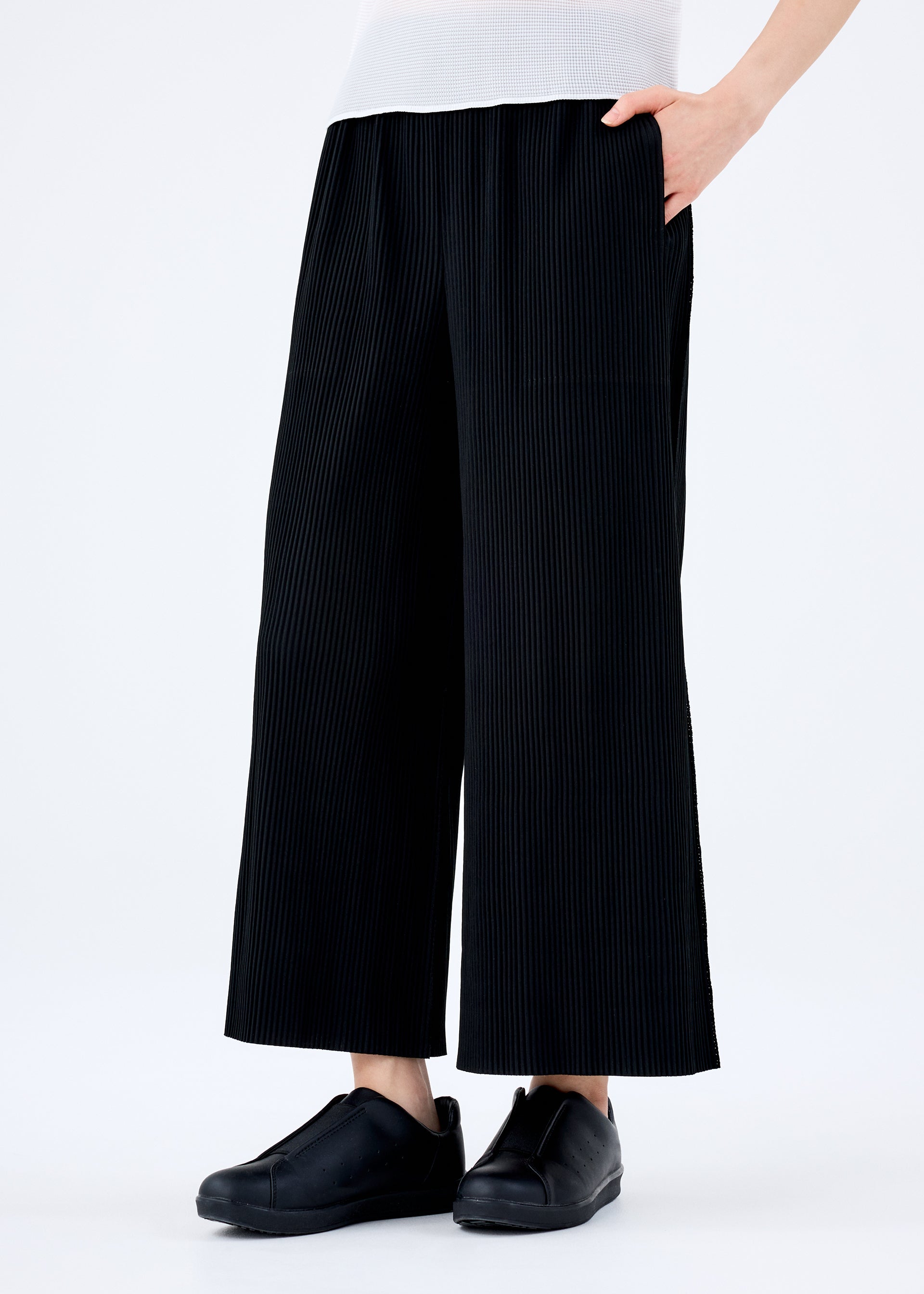Zoomed view A person wears the FINE KNIT PLEATS BLACK pants by me ISSEY MIYAKE with their right hand in a pocket, paired with black slip-on shoes and a white top, standing against a plain white background.