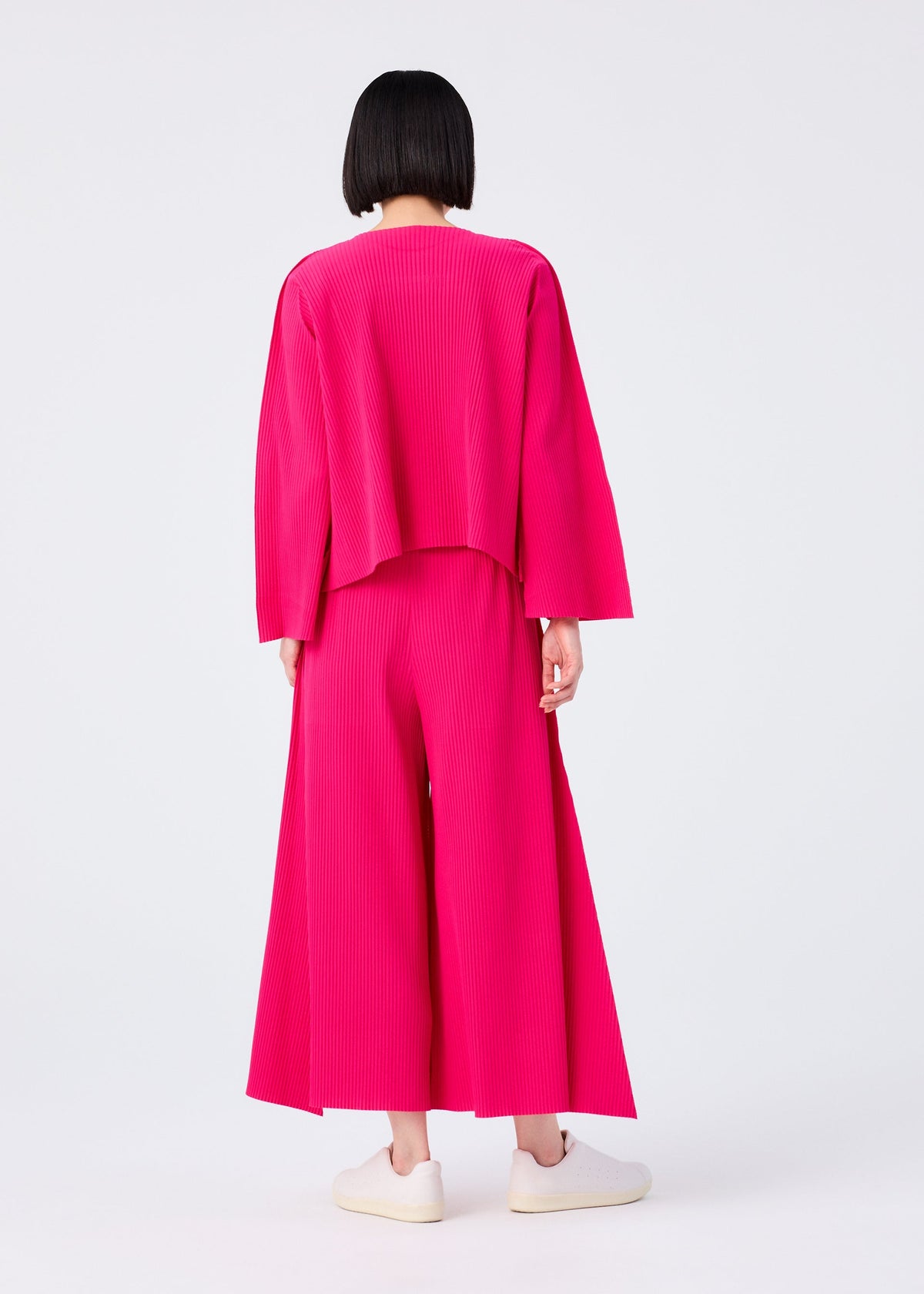 A person with a short black bob stands facing away, wearing the FLARE PLEATS set—loose-fitting, long-sleeved bright pink top and matching wide flared pants—from me ISSEY MIYAKE, with white slip-on shoes against a plain white background. view 7 of 8
