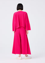 A person with a short black bob stands facing away, wearing the FLARE PLEATS set—loose-fitting, long-sleeved bright pink top and matching wide flared pants—from me ISSEY MIYAKE, with white slip-on shoes against a plain white background.