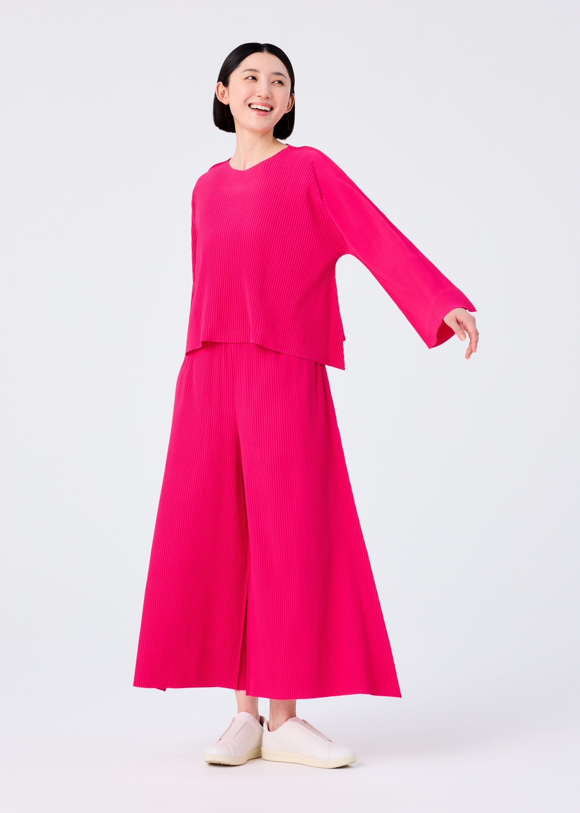 Zoomed view A person with short black hair smiles, modeling the FLARE PLEATS set by me ISSEY MIYAKE—a bright pink long-sleeve top and wide flared pants—paired with white slip-on shoes against a plain white background.