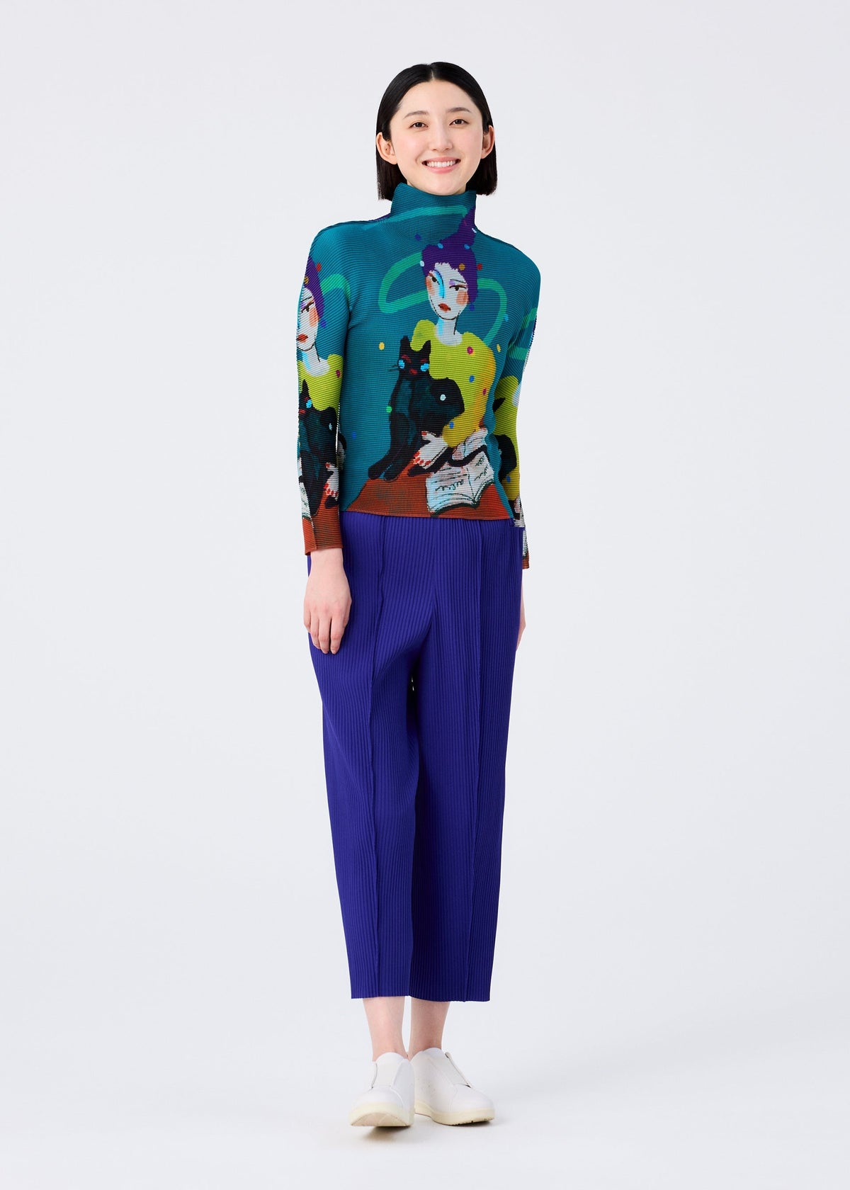 A woman smiles against a plain white background, wearing the FINE KNIT PLEATS COLOR 1 sweater by me ISSEY MIYAKE with artistic faces and designs, paired with loose straight pants and white slip-on shoes. view 7 of 8