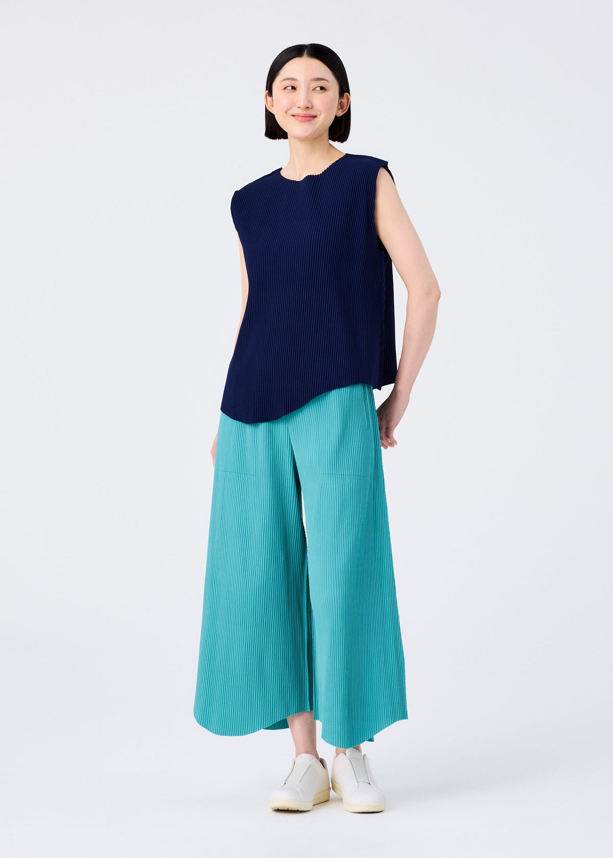 A woman with short black hair smiles in a sleeveless navy top and aqua FINE KNIT PLEATS BOTTOM 1 pants by me ISSEY MIYAKE, paired with white shoes against a plain white background. view 5 of 6